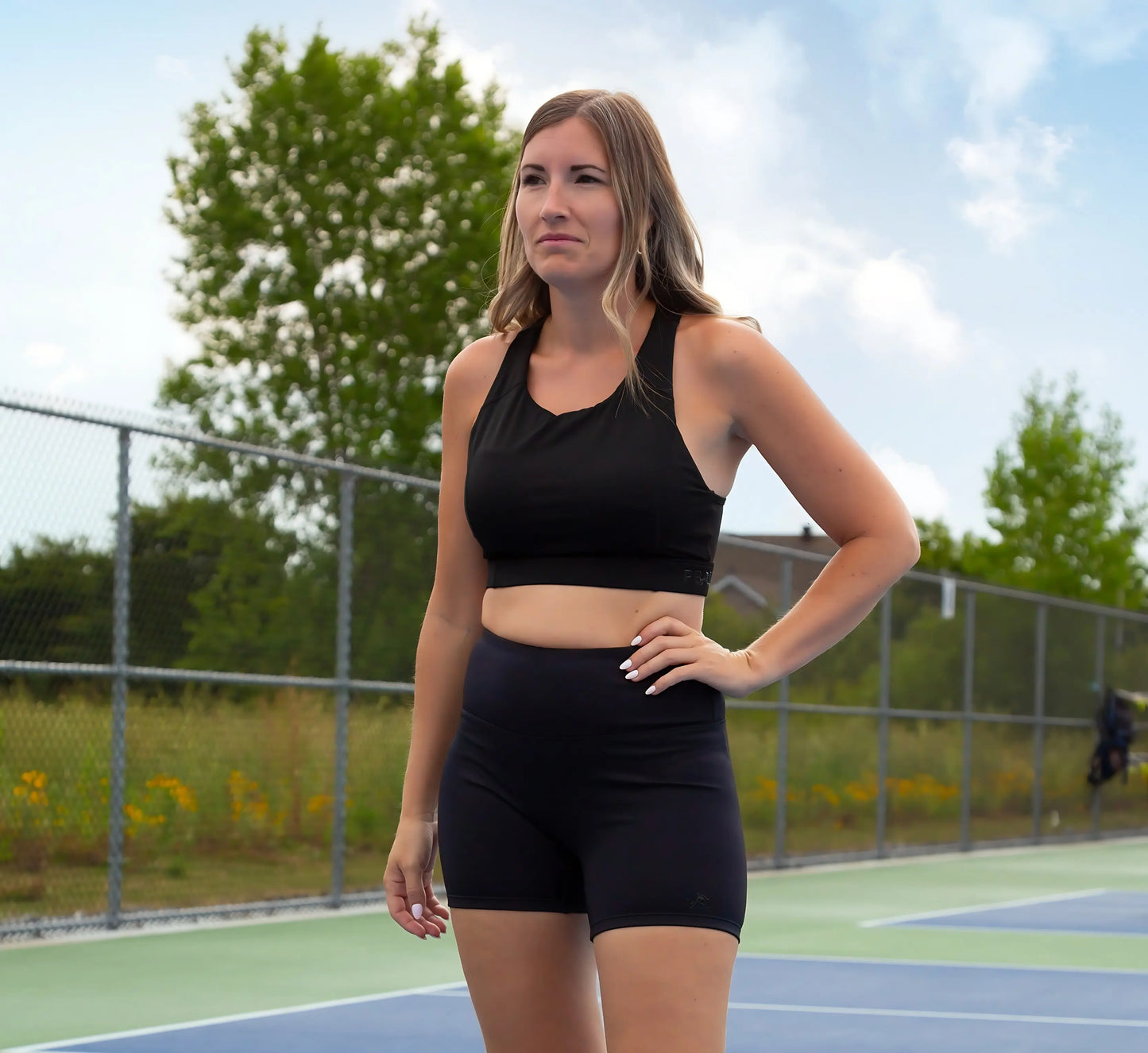 Woman in black athletic wear standing on a pickleball court with trees and sky in the background