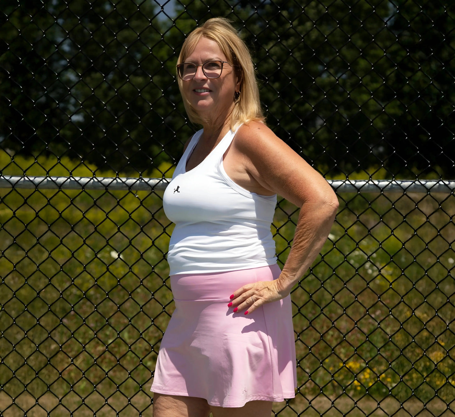 Woman in a White tank top and pink skirt standing in front of a chain-link fence Blanc