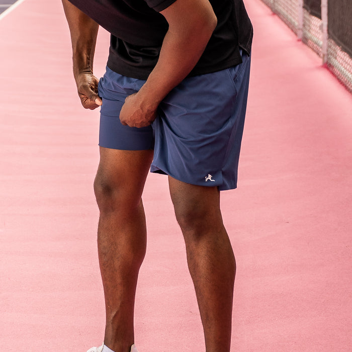 A close-up of a person wearing blue PBALL pickleball shorts and a black shirt on a pink court, with the shorts showing a small white logo near the hem.