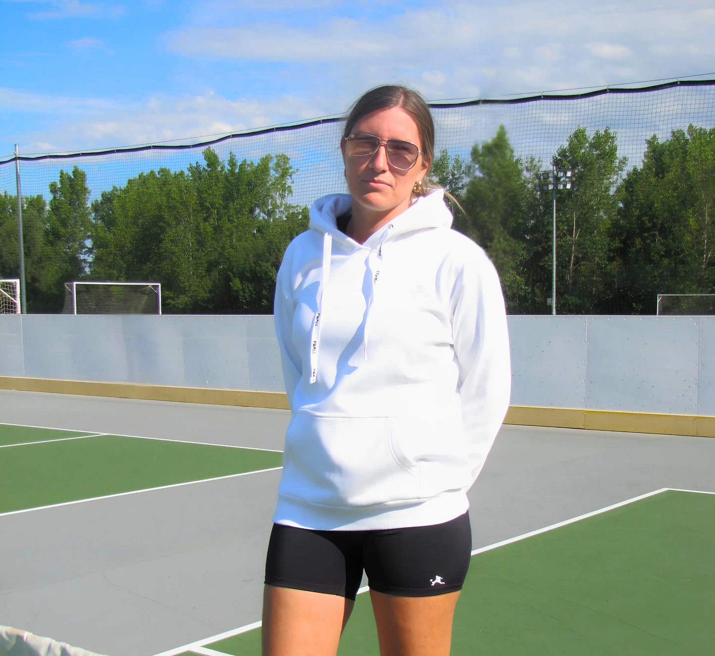 Woman wearing a White hoodie and shorts on a pickleball court with trees in the background Blanc