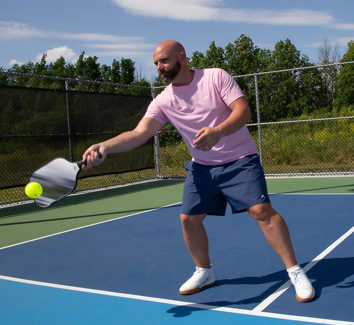 Man wearing Blue shorts playing pickleball on a court with trees in the background Bleu