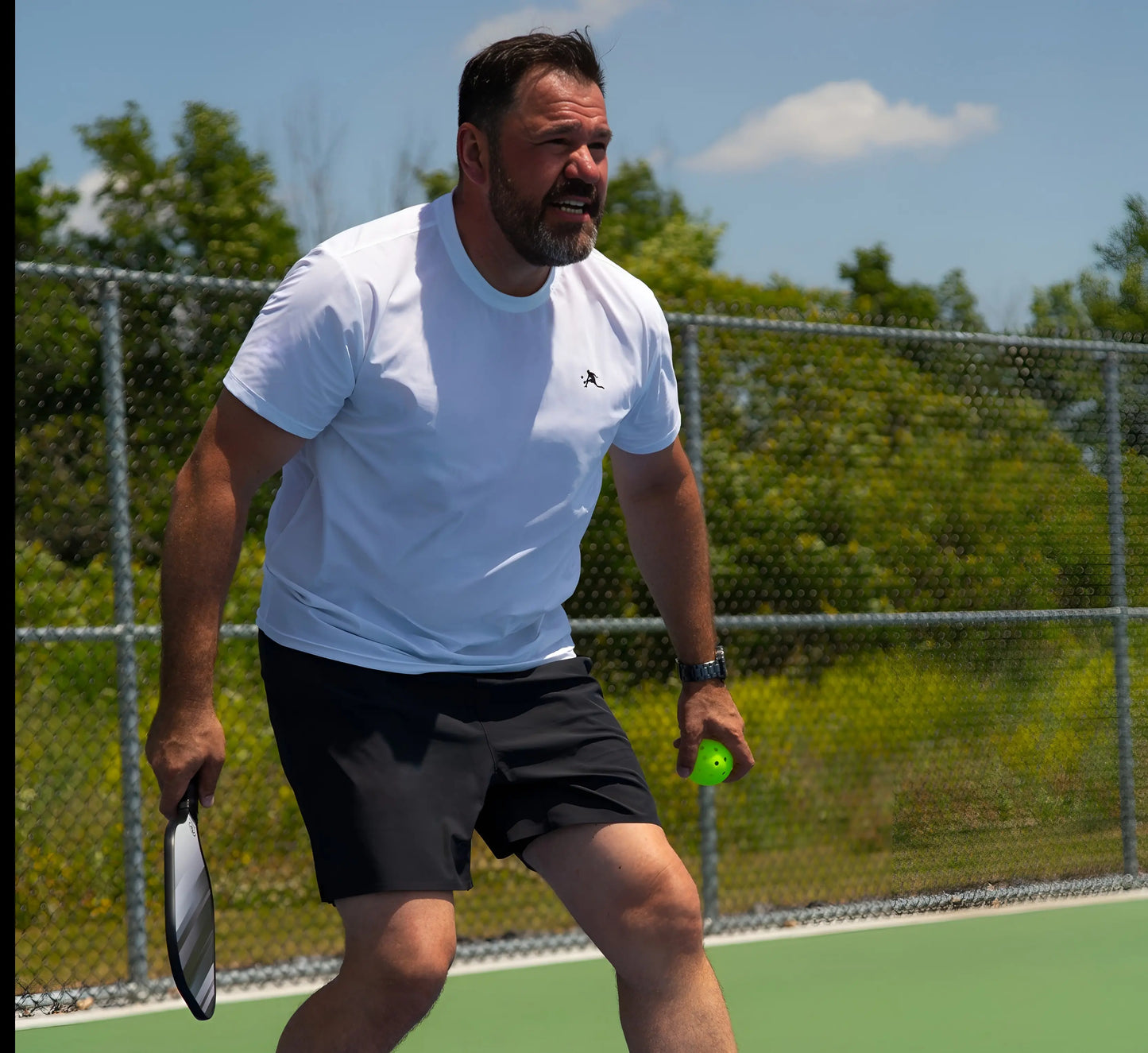 Man wearing Black shorts playing pickleball on a court with trees and a fence in the background Noir