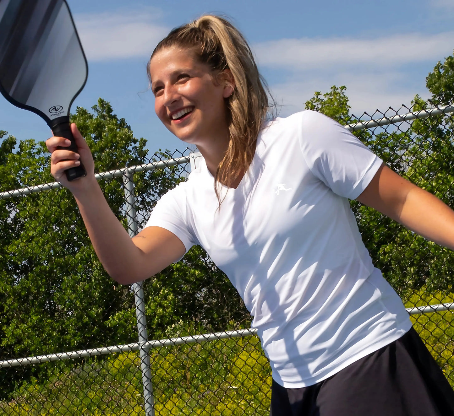 Woman wearing a White t-shirt holding a pickleball paddle on a court with trees and sky in the background Blanc