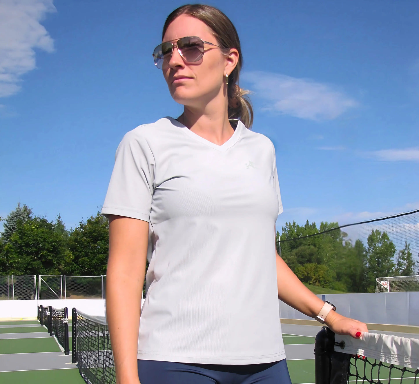 Woman standing on a pickleball court wearing sunglasses and a light Grey t-shirt Gris
