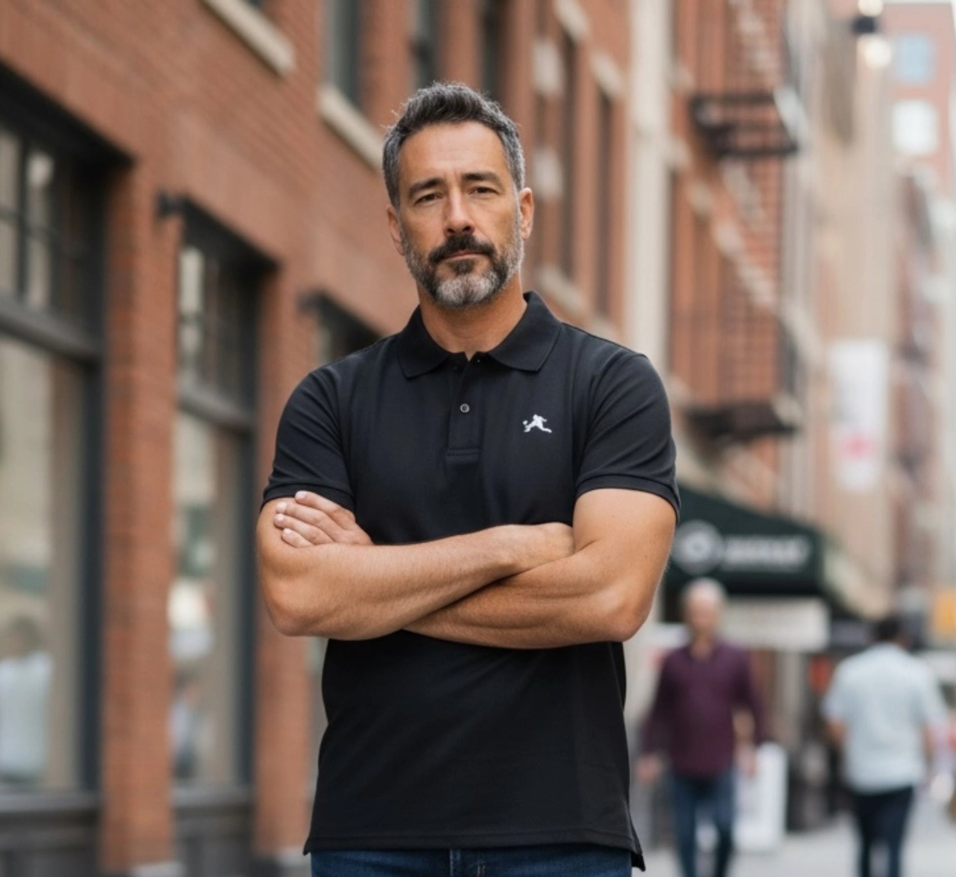 Man wearing a black polo shirt with a logo standing on a city street.