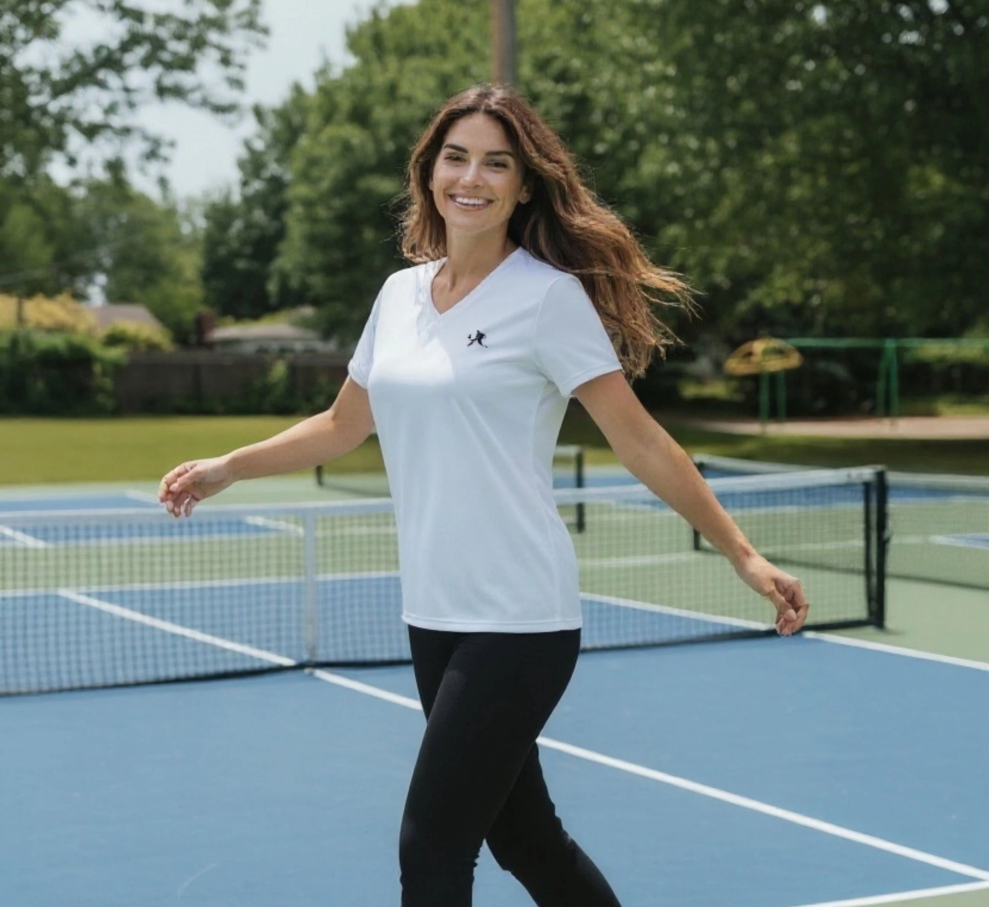 Woman standing on a pickleball court wearing a white t-shirt with a logo.