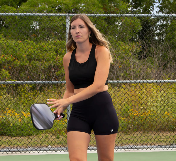 Woman in black athletic wear holding a pickleball paddle on a court with a chain-link fence and greenery in the background.
