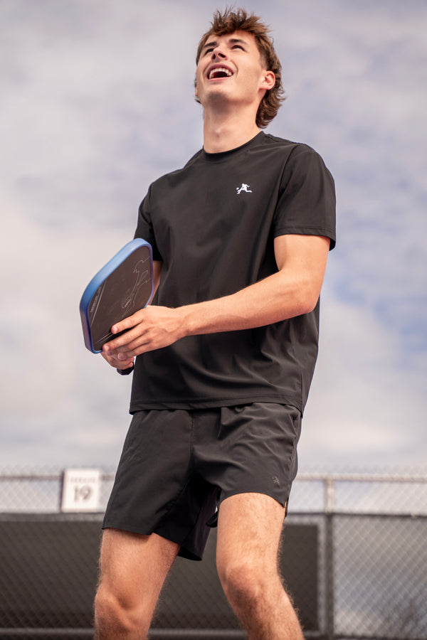 A man laughing on an outdoor court wearing a black PBALL t-shirt and black pickleball shorts, holding a paddle with a blue and black grip.