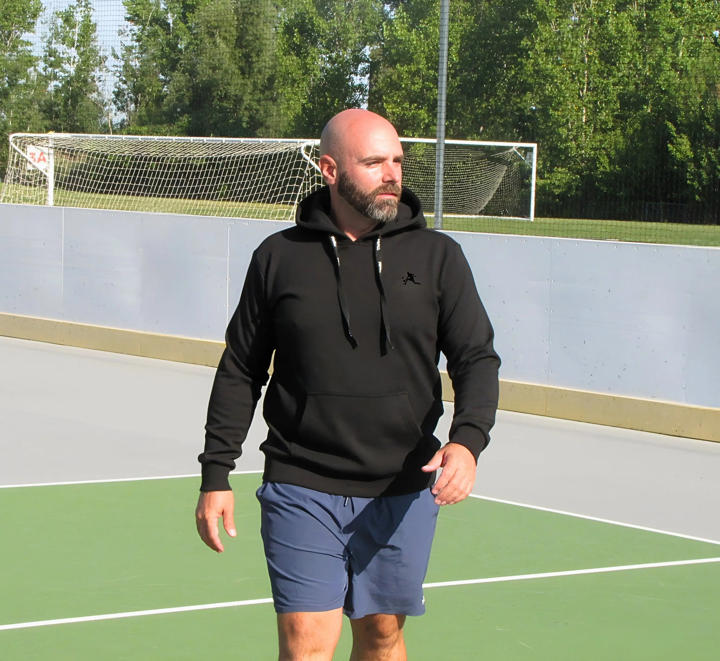 Man wearing a Black hoodie and shorts on an outdoor court with trees in the background Noir