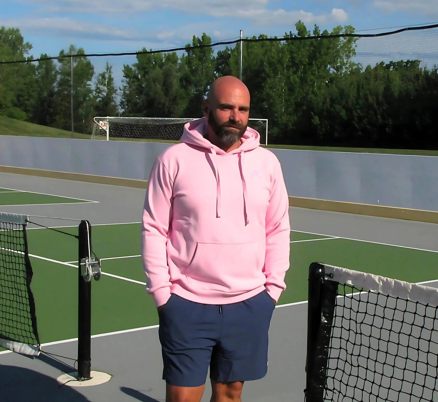 Man in a Pink hoodie standing on a pickleball court with trees in the background Rose