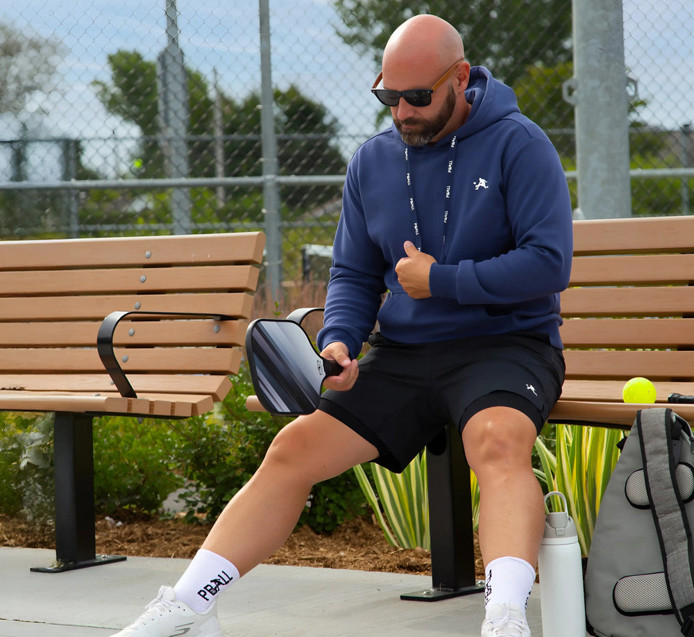Homme assis sur un banc, tenant une raquette de pickleball, portant un Hoodie et un short Bleu. Blue