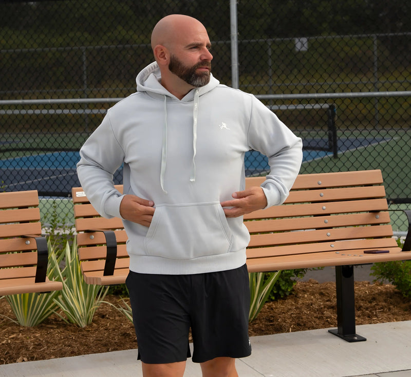 Homme portant un sweat à capuche Gris et un short noir, debout sur un trottoir avec des bancs et une clôture en arrière-plan. Grey