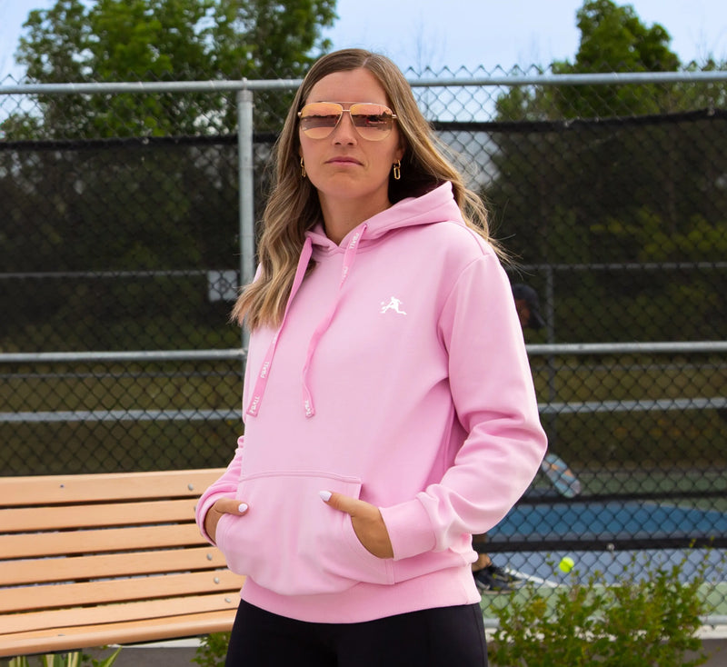 Femme portant un sweat à capuche Rose, debout sur un terrain de pickleball avec une raquette et une balle. Pink