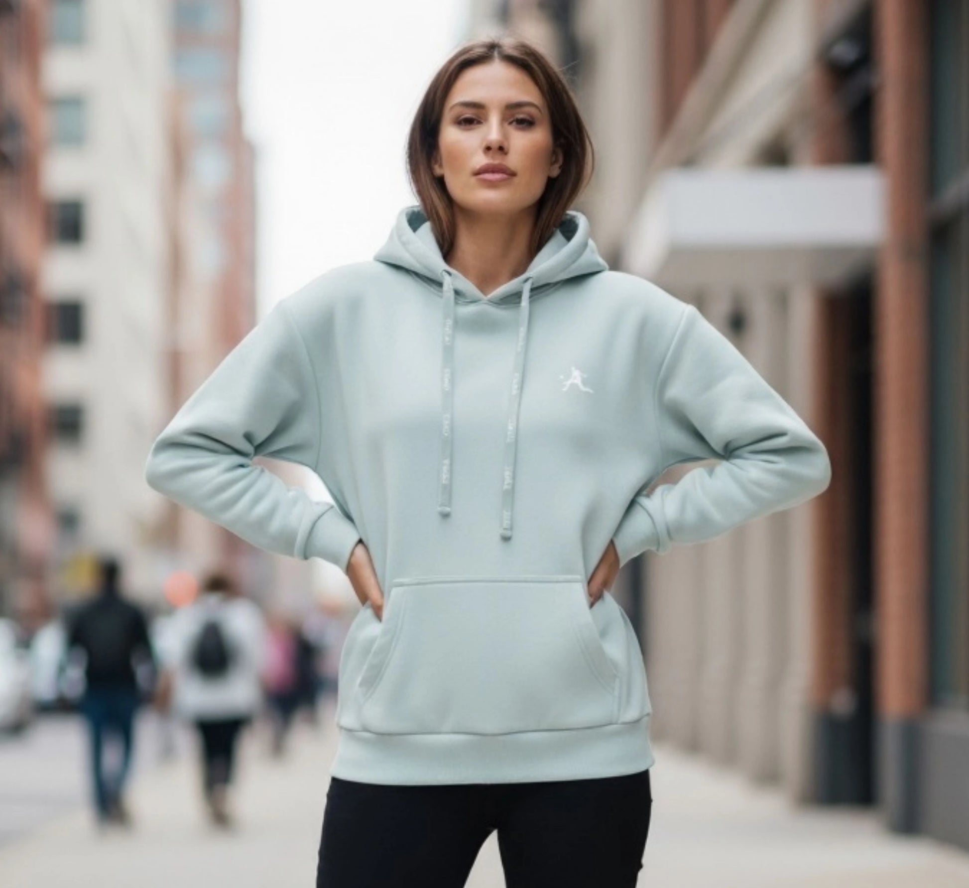 Woman wearing a light blue hoodie with a brand logo on a city street.