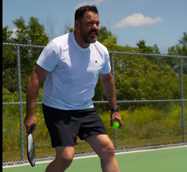 Man wearing Black shorts playing pickleball on a court with trees and a fence in the background Noir