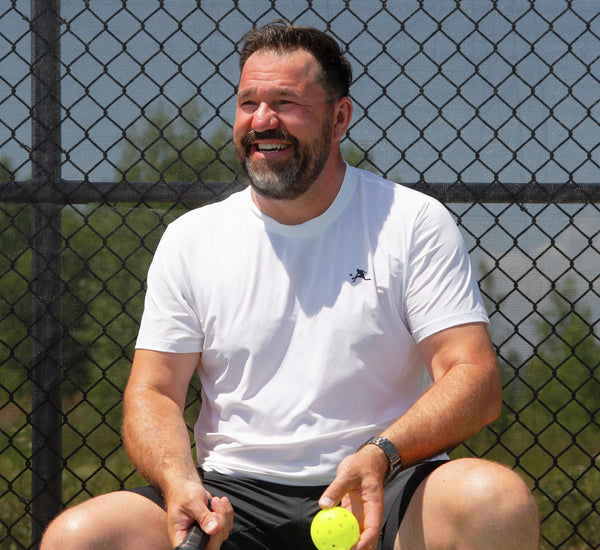 Man wearing a White t-shirt sitting on a pickleball court holding a paddle and ball, with a chain-link fence in the background Blanc