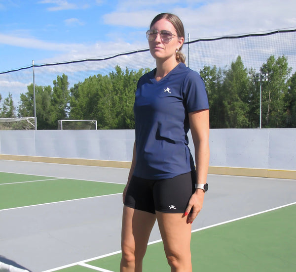Woman in a Blue t-shirt standing on a pickleball court with trees in the background Bleu