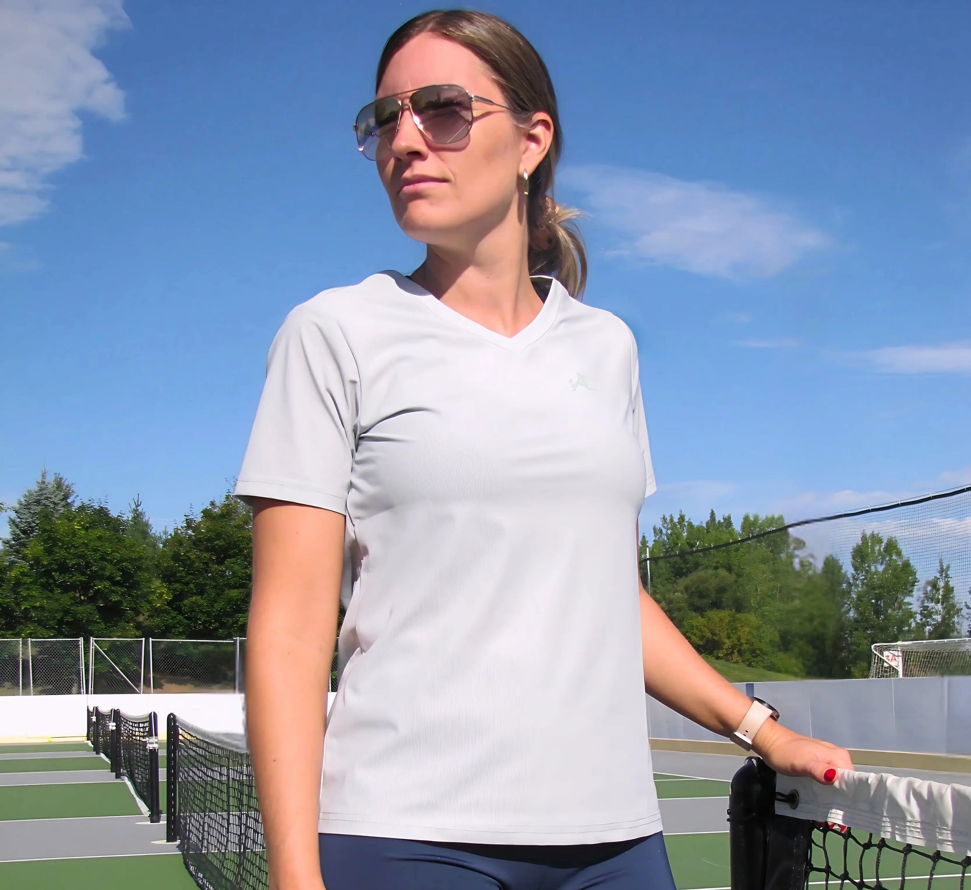 Woman standing on a pickleball court wearing sunglasses and a light Grey t-shirt Gris