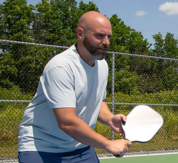 Man wearing a Grey t-shirt playing pickleball on a court with trees in the background Gris